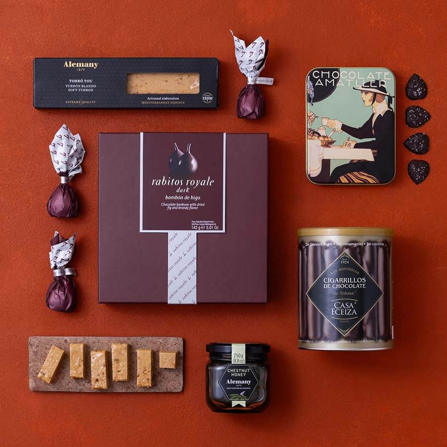 A flat-lay arrangement of gourmet Spanish snacks and chocolates on a textured terracotta background.