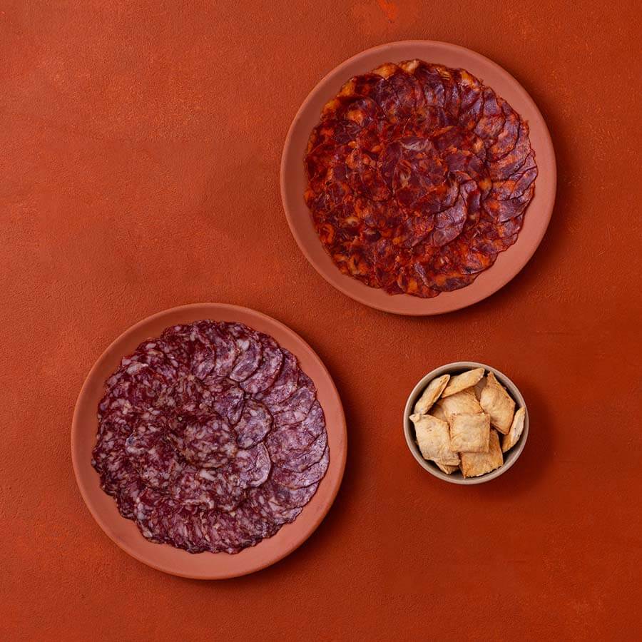 Two round terracotta plates filled with sliced cured meats, one reddish salami and one darker chorizo-style sausage, arranged on a warm orange-brown background with a small bowl of square crackers.