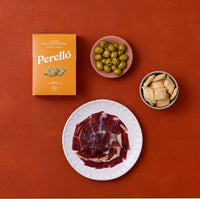 A styled flat-lay of Perelló Classic Olive Oil Crackers on a terracotta background, featuring the yellow-orange product box, a white plate with sliced cured meat, a pink bowl of green olives, and a gray bowl of flatbreads.
