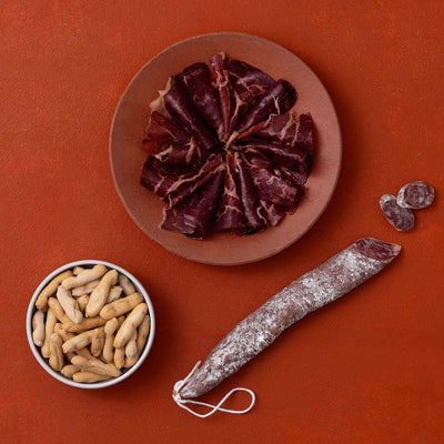 A styled flat-lay of cured meats and picos camperos on a terracotta background.