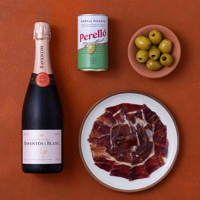 A styled flat-lay of a bottle of Raventos Blanc sparkling wine, a can of Perello Gordal Picante, a small bowl of green olives, and a plate of sliced cured ham on a terracotta background.