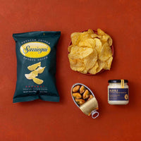 A styled flat-lay of packaged snacks on a terracotta background: a dark teal bag of Sarriequi potato chips, a red bowl filled with chips, an open can of mussels, and a jar of Aioli sauce.