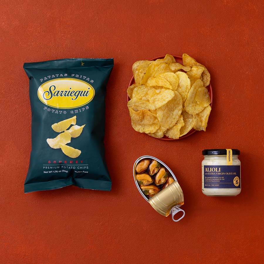 A styled flat-lay of packaged snacks on a terracotta background: a dark teal bag of Sarriequi potato chips, a red bowl filled with chips, an open can of mussels, and a jar of Aioli sauce.