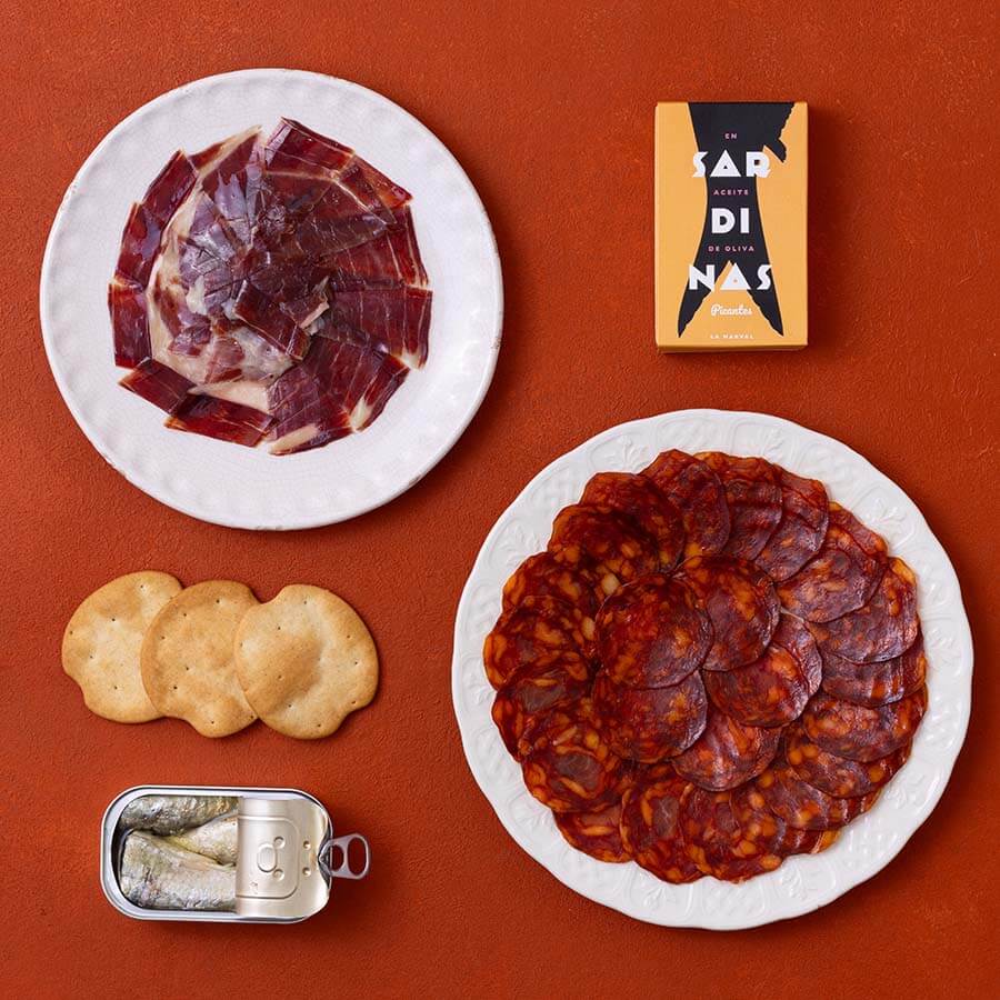 A styled flat-lay of Spanish charcuterie items on a terracotta background: a white plate with sliced cured ham, a white plate with sliced chorizo, a small open tin of sardines and three round crackers.