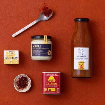 A styled flat-lay of multiple food products on a terracotta background: a large bottle of fumet, a jar of alioli, a box of saffron, a can of La Chinata paprika, a small dish with saffron, and a spoon with paprika. 