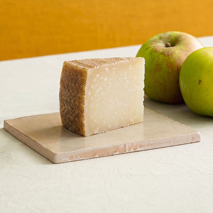 A rectangular wedge of Manchego cheese with a textured brown rind sits on a square beige stone board, with two green apples blurred in the background against a warm yellow wall.