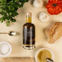 Bottle of garlic and parsley infused olive oil laid flat on a table and surrounded by bread, salt, parsley, tomato and garlic.
