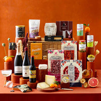 An assortment of Spanish products arranged on terracotta prop with a warm terracotta backgrounds. Visible products include sliced charcuterie, wines, cheeses and olives. 
