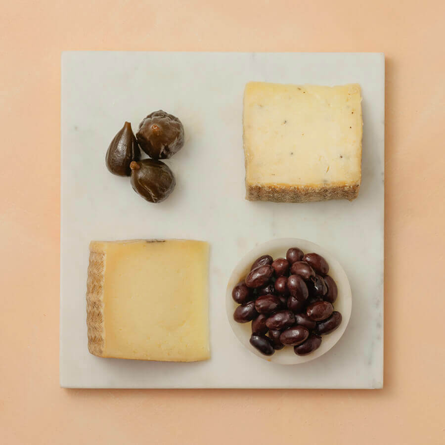 Two blocks of cheese and a bowl of olives on a marble board with a pink background