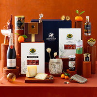 A styled gourmet gift hamper featuring an assortment of Spanish products including cheese, charcuterie, sparkling wine and snacks, arranged on terracotta pedestals against a matching terracotta background.