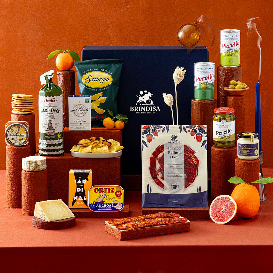 A Spanish selection of products arranged on terracotta pedestals against a warm terracotta background. Visible products include ham, chorizo, crisps, olives and alioli. 