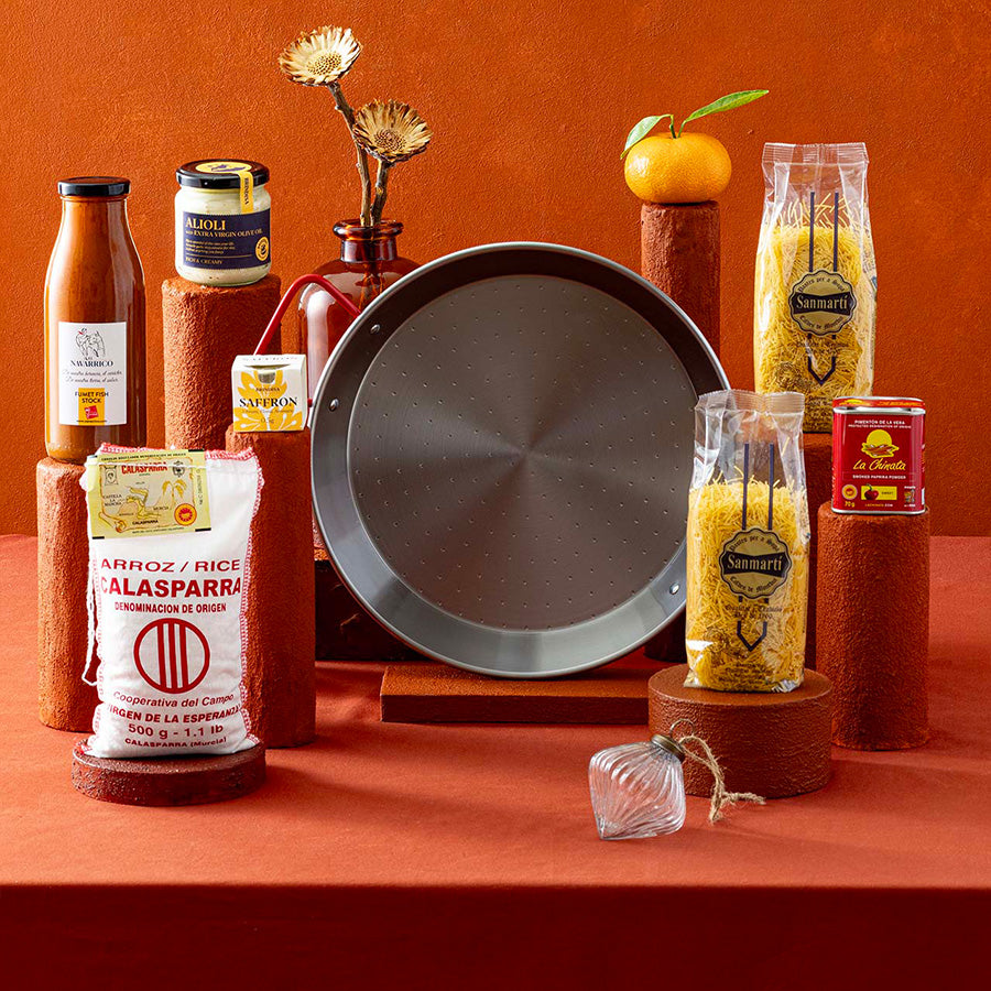 A styled gift hamper featuring a large round paella pan as the central item, surrounded by various Spanish food ingredients and condiments used for paella and fideus recipes, on wooden pedestals against a terracotta background.