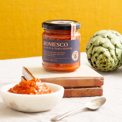 Jar of Brindisa Romesco sauce beside a fresh artichoke, ceramic tiles, and a bowl of rich, red romesco with a spoon