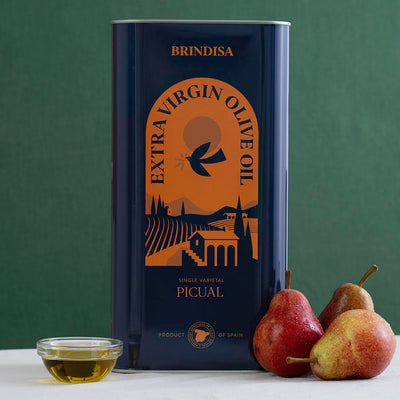 A tall tin of Brindisa Extra Virgin Olive Oil Picual stands on a white surface against a dark green background, with a small glass bowl of olive oil and three pears placed in front of the tin.