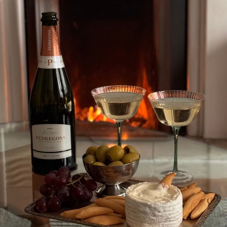A bottle of Cava beside a soft cheese and picos de pan, with two full wine glasses set in front of a lit fireplace.