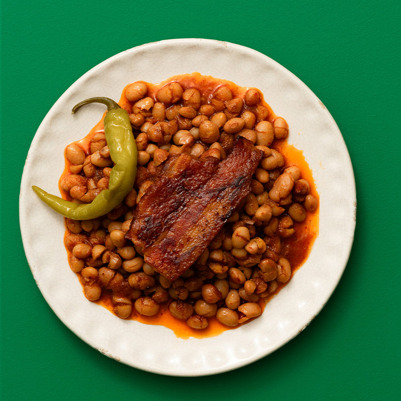 A top-down view of a white plate containing baked beans in a reddish-orange sauce with a piece of crispy bacon and a green chili pepper, set against a solid green background.