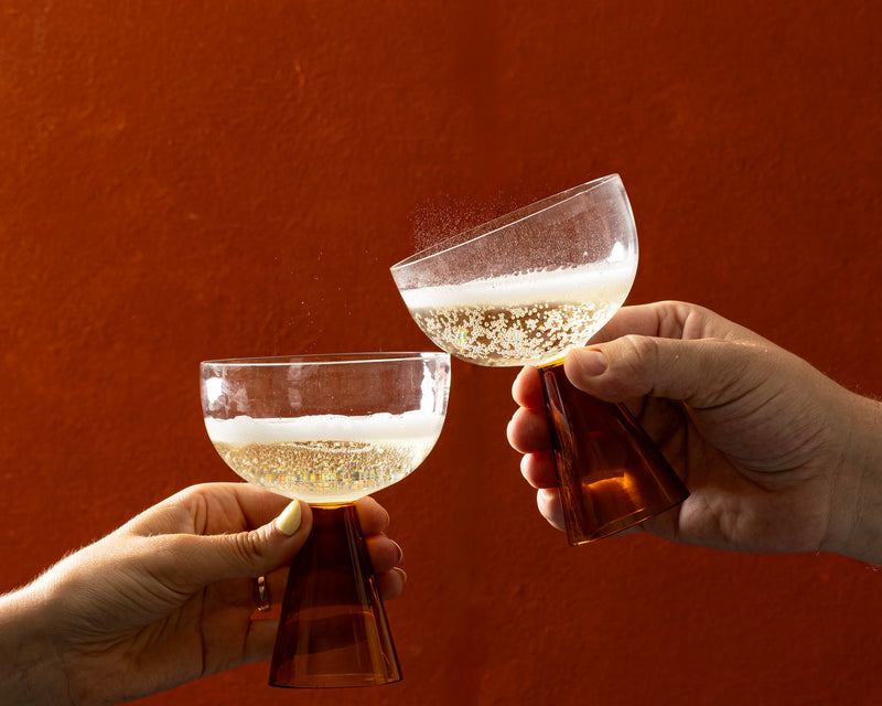 Two hands holding up glasses in a toast, clinking together against a textured rust-orange background.