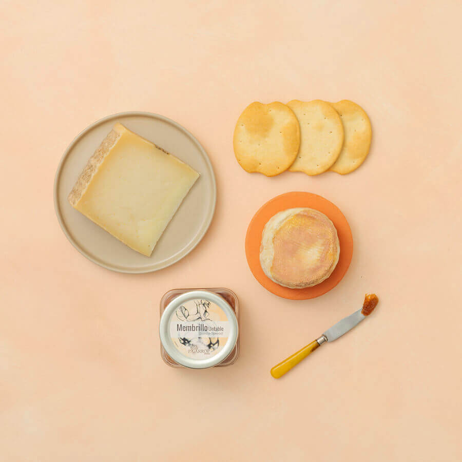 Torta-style cheese, manchego cheese, ships crackers and a knife with me, with membrillo on a peach background