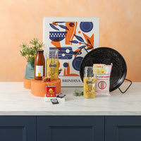 Tabletop display with decorative items including a bottle of fumet, a paella pan, Christmas decorations such as candles, pasta packs and rice on a peach-colored wall.