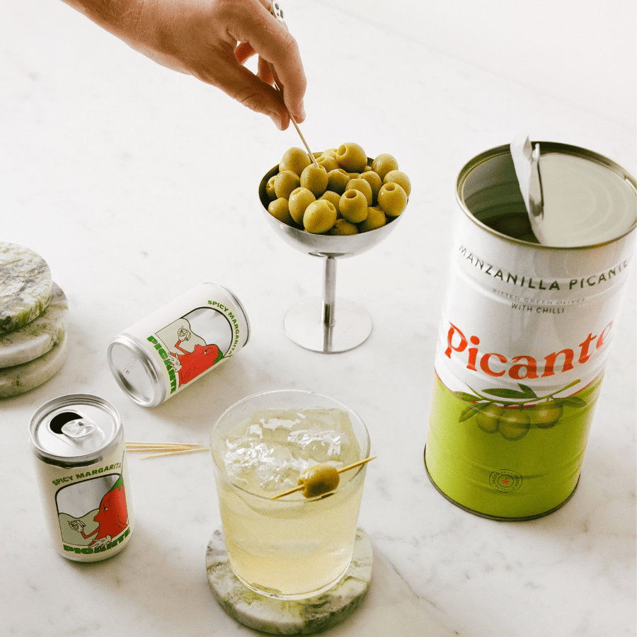 Cocktail with olives, Picante cocktail cans, and a bowl of olives on a marble surface.